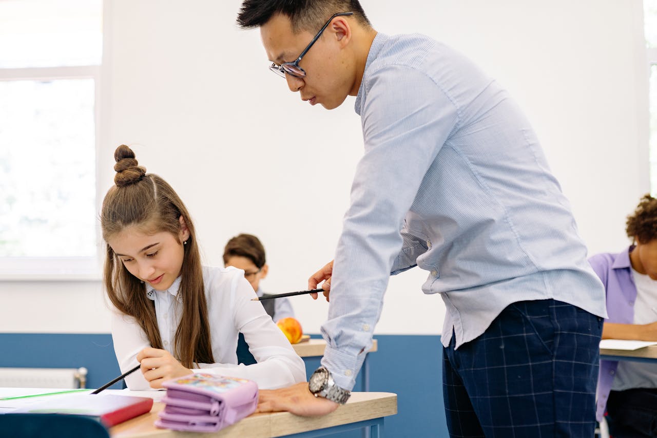 Asian male teacher assisting a young caucasian girl with her studies in a classroom setting.